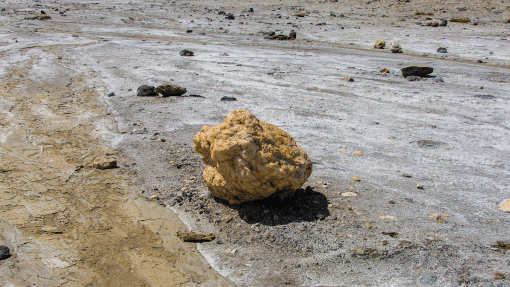 Pedra grande e solitária em área seca e rachada do deserto, cercada por outras menores ao fundo.