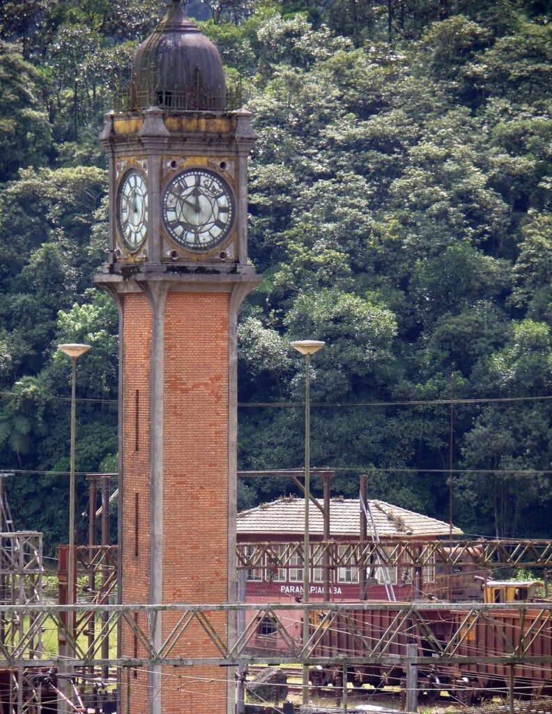 Torre histórica com relógios de época e letreiro de Paranapiacaba ao fundo, entre trilhos e vagões abandonados.