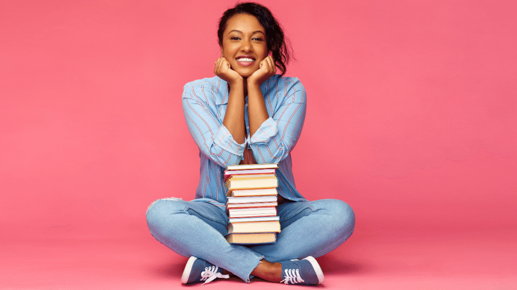 Mulher sorridente sentada no chão com uma pilha de livros no colo